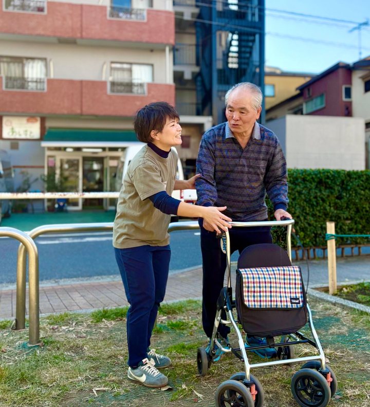 公園で歩行器を使って歩く男性に寄り添うスタッフ